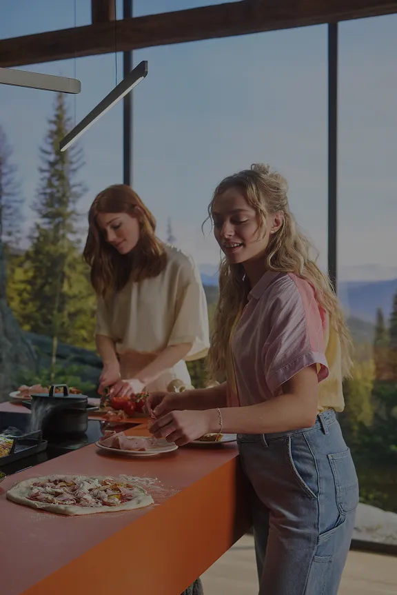 Mann und zwei Frauen bereiten in einer modernen Küche mit Bergblick Pizza und andere Speisen zu. Er hält eine Pizza, sie schneiden Gemüse.